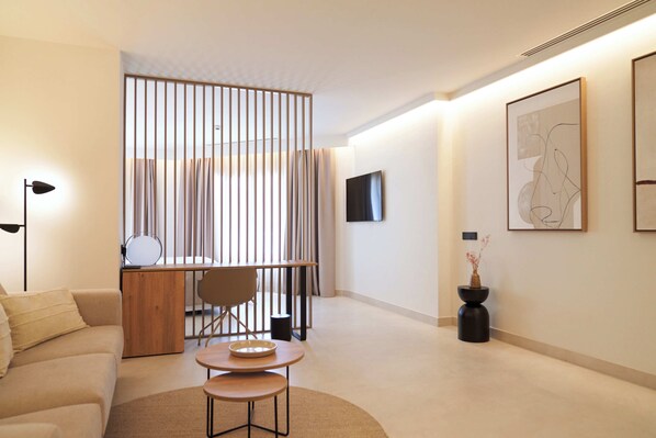 Una habitación de hotel moderna con sofá, una mesa de comedor, televisor montado en la pared y un jarrón decorativo.