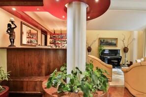 Un hall d’entrée avec un comptoir de réception en bois, un coin bar avec des étagères remplies de bouteilles, un piano et plusieurs plantes en pot.