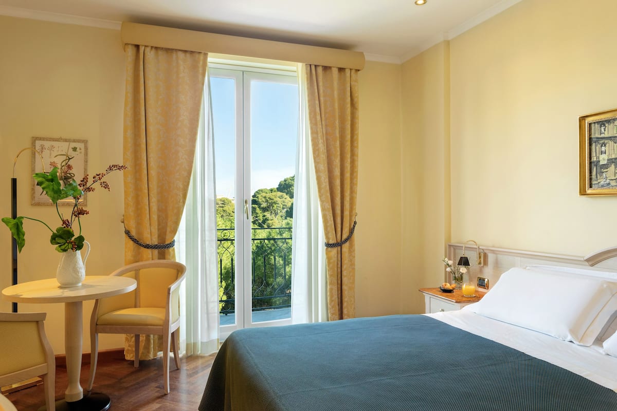 Une chambre d’hôtel avec un lit, une chaise, une petite table sur laquelle est posé un vase de fleurs, et une fenêtre avec des rideaux.