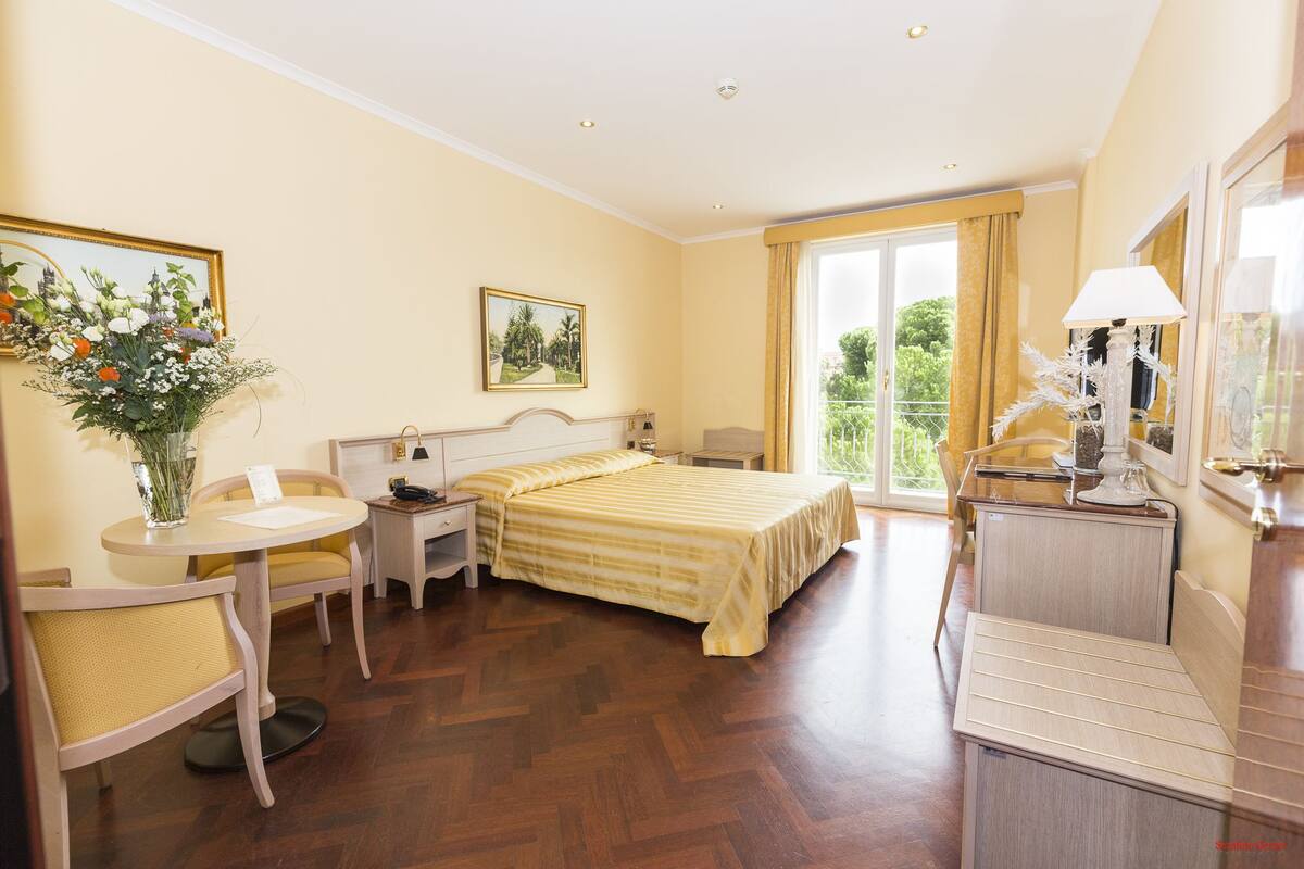 Une chambre d’hôtel comprenant un lit, un bureau, une chaise, une table de chevet, une lampe et une fenêtre donnant sur un paysage verdoyant.