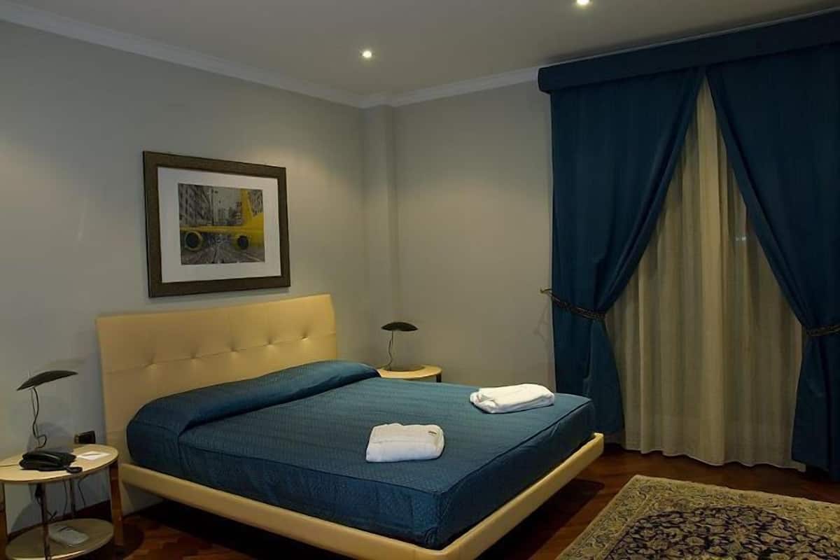 Une chambre d’hôtel avec un lit, une table de chevet, une lampe, un tableau encadré au mur et une fenêtre avec des rideaux.