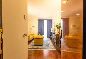 Een hotelkamer met een houten vloer, een gele fauteuil, een blauw vloerkleed, een dressoir en een raam met blauwe gordijnen.