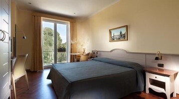 Camera d'albergo con un letto, una scrivania, una sedia, un comodino e una finestra vista verde.