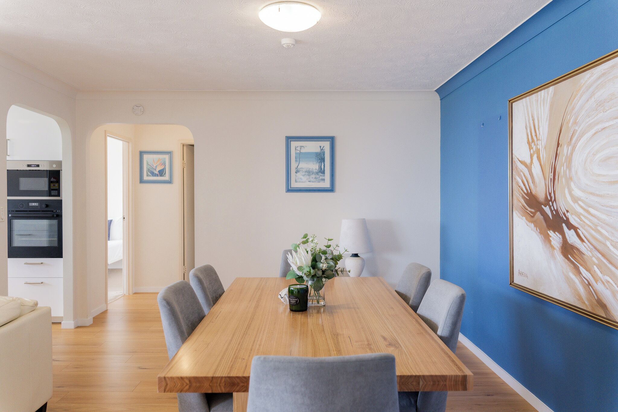 A dining area with a wooden table, six chairs, a vase with flowers, and two framed pictures on the wall.