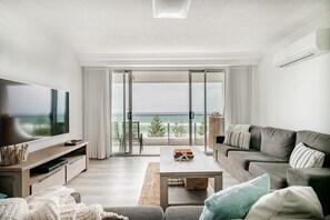 Una sala de estar moderna con un televisor de pantalla plana grande, un sofá gris, una mesa de centro y vistas al océano a través de puertas correderas de vidrio.