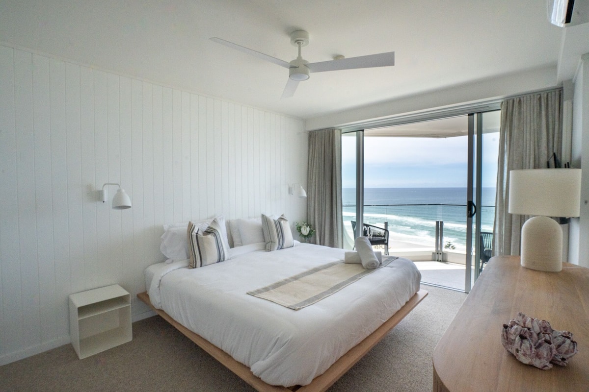 Habitación de hotel con una cama grande, un ventilador de techo, vista al océano y una mesita de noche de madera.