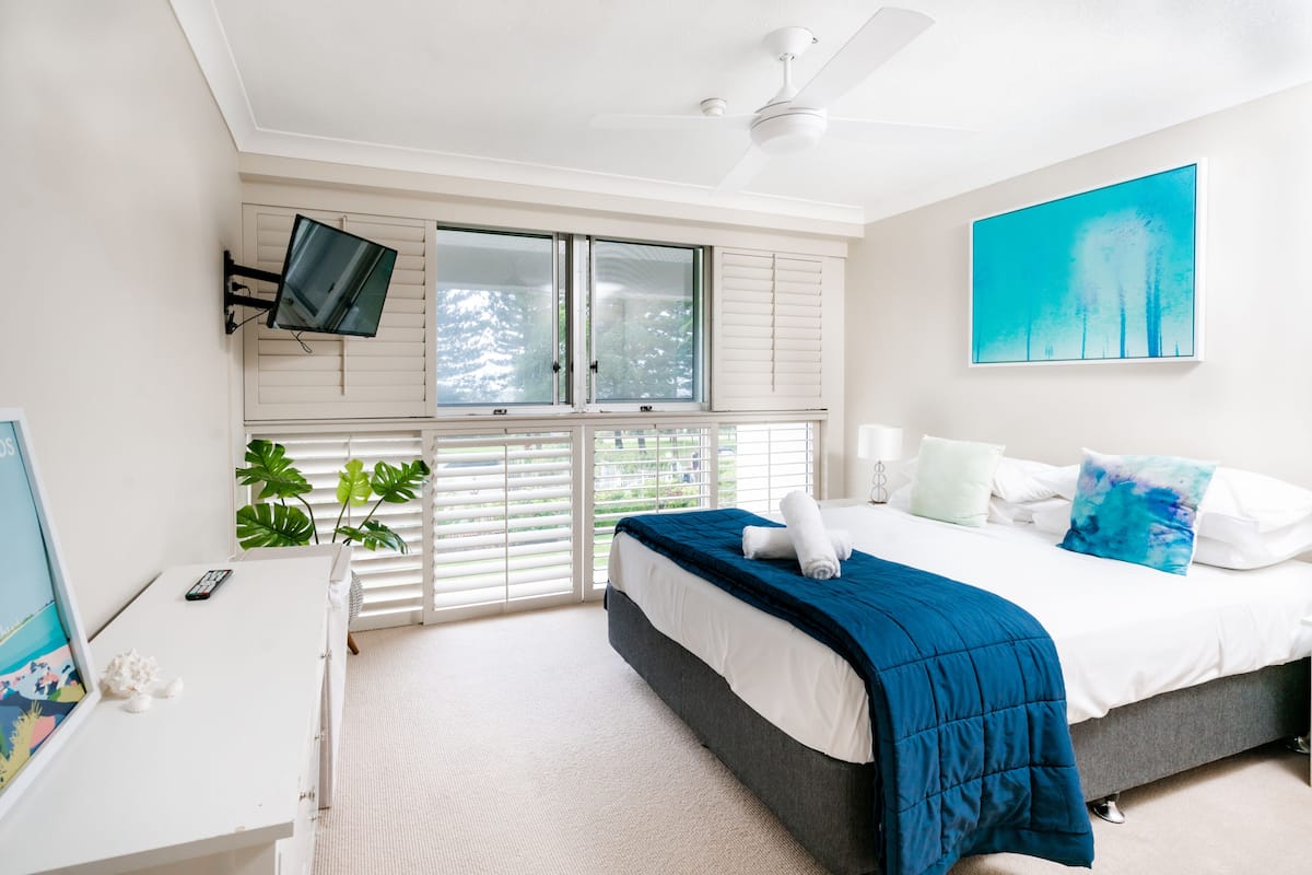 Un dormitorio con una cama, un televisor montado en la pared, un ventilador de techo y una ventana con contraventanas.