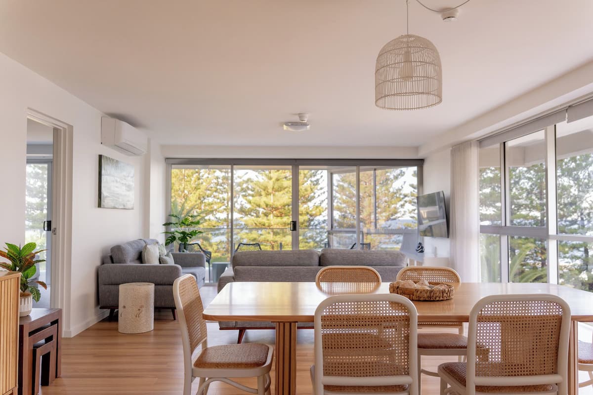 Una sala de estar moderna con una mesa de comedor de madera, sillas de mimbre, un sofá gris y ventanas amplias que ofrecen vistas a árboles.