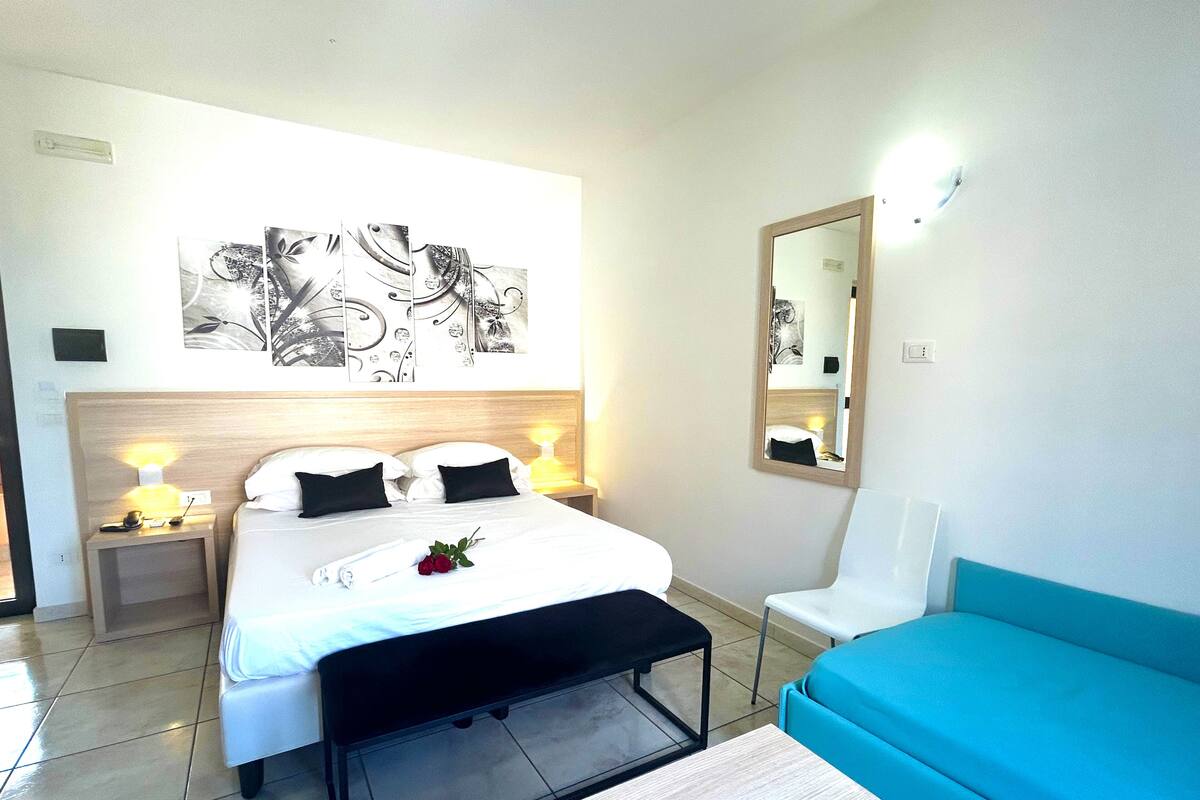 A hotel room with a bed, bedside lamps, a mirror, a blue sofa, and a wall with abstract art.