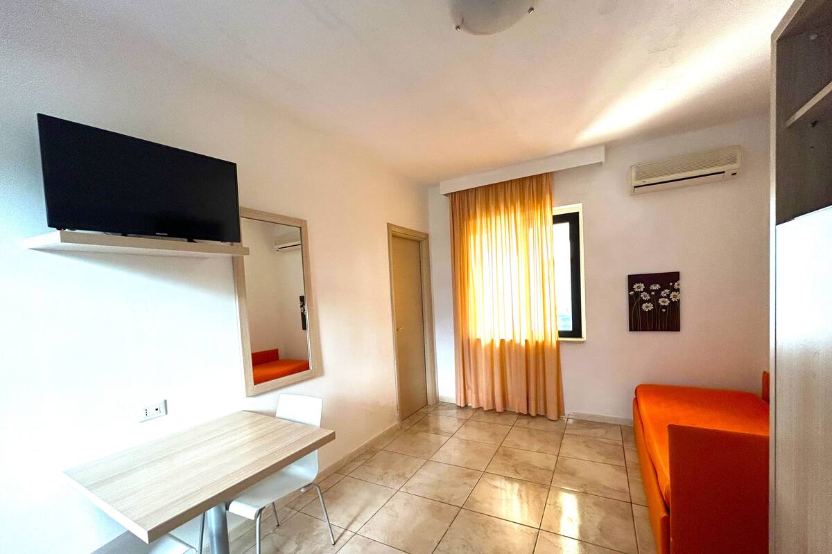 A room with a flat-screen TV mounted on the wall, a wooden desk, a chair, a bed with an orange cover, a window with curtains, and a wall-mounted air conditioner.