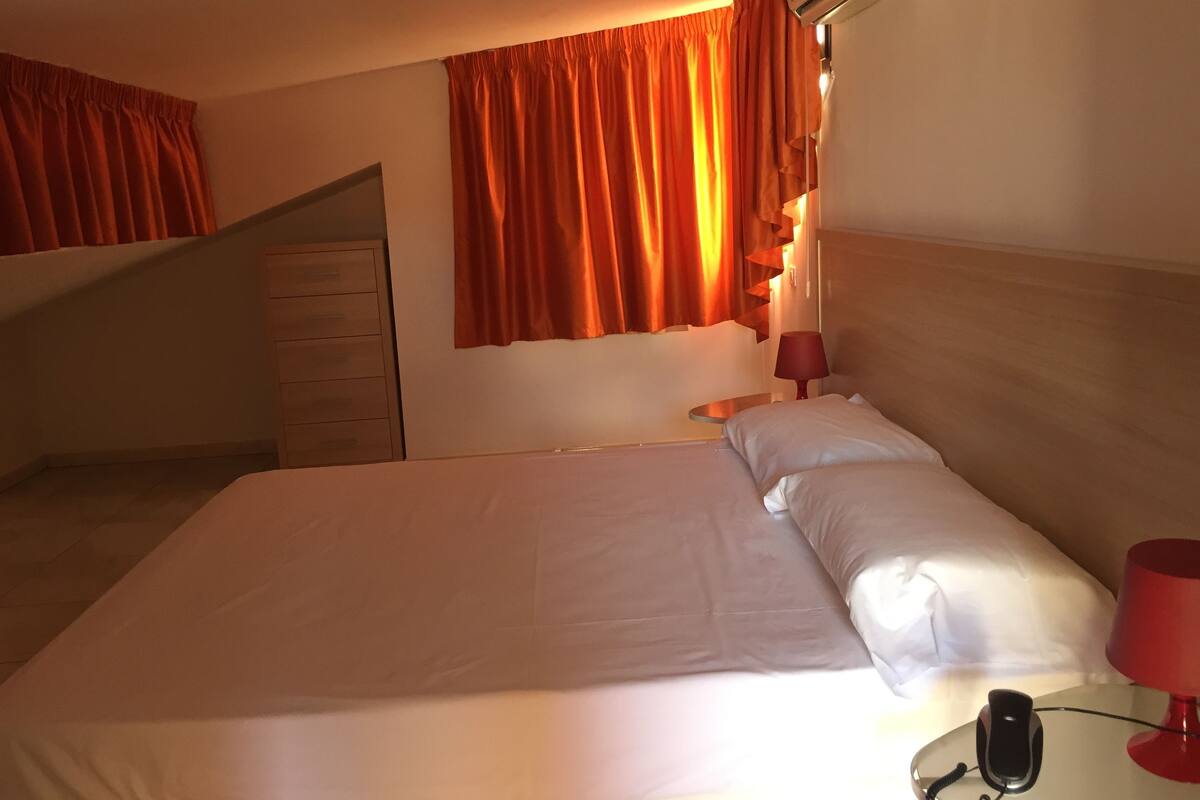 A hotel room with a bed, a bedside table with a lamp, a telephone, and a wall-mounted air conditioner.