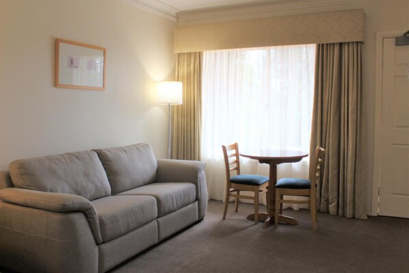Quarto de hotel com um sofá bege, uma mesa redonda pequena com duas cadeiras, um abajur e uma janela com cortinas.