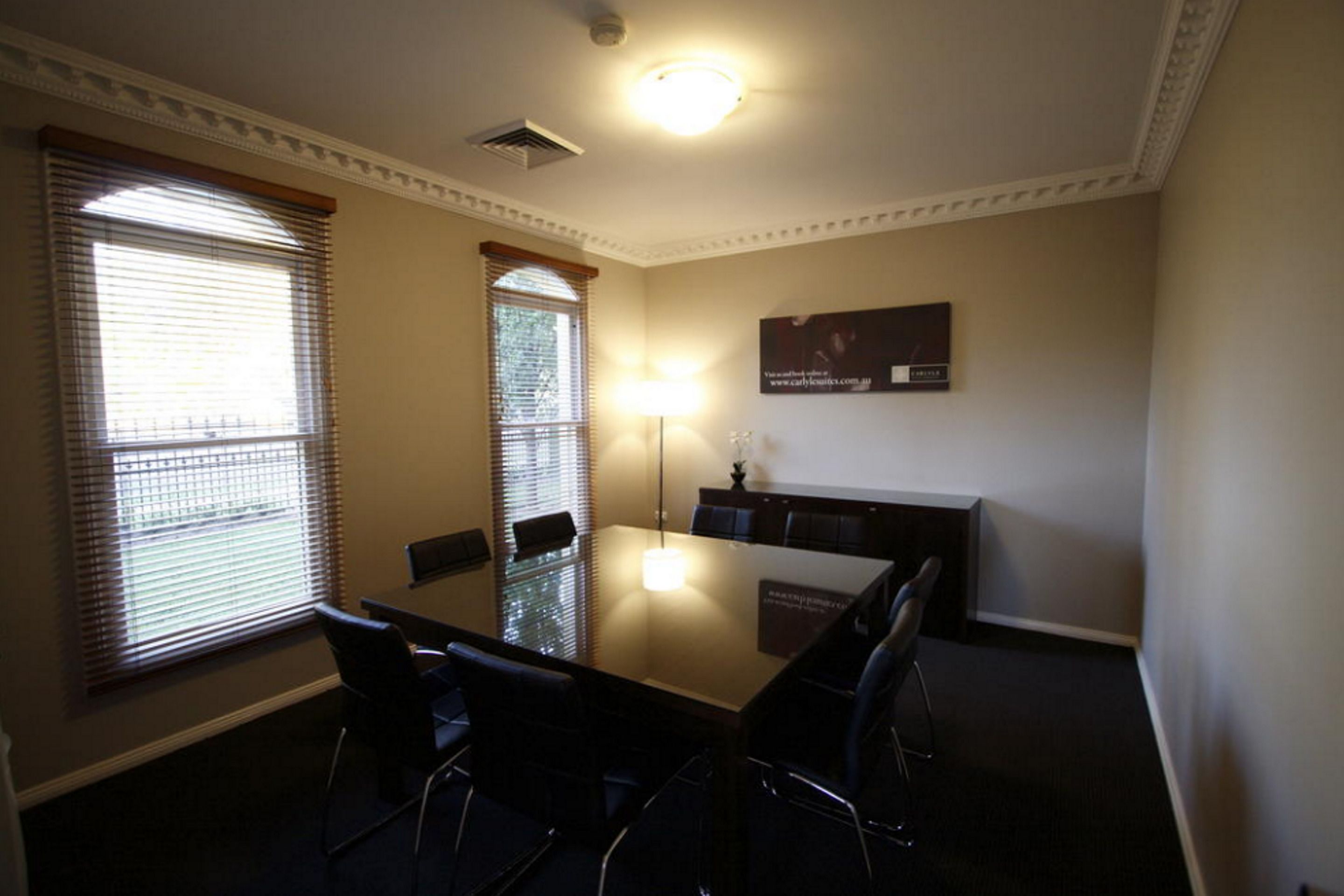 A conference room with a glass table, six chairs, two windows with blinds, a ceiling light, and a sign on the wall.