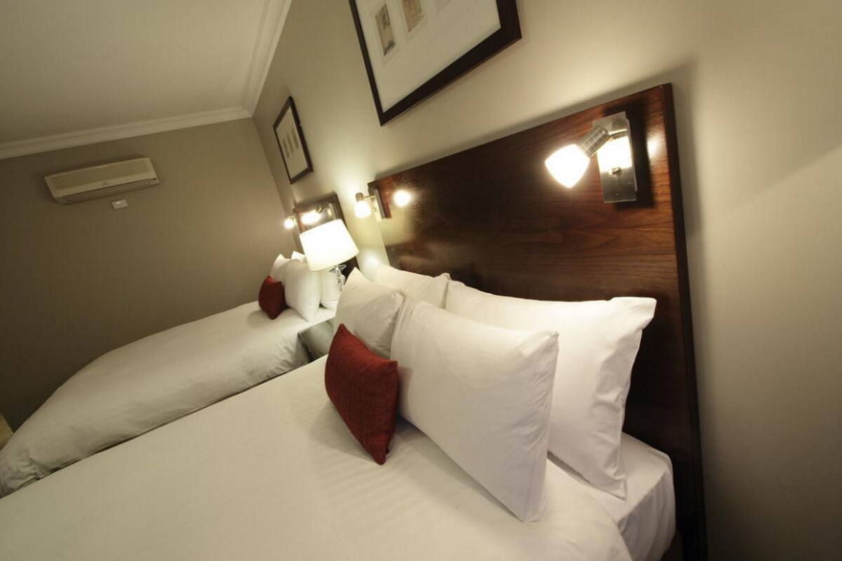 A hotel room with two beds, each with white bedding and red pillows. There are wall-mounted lamps and a framed picture above the beds.