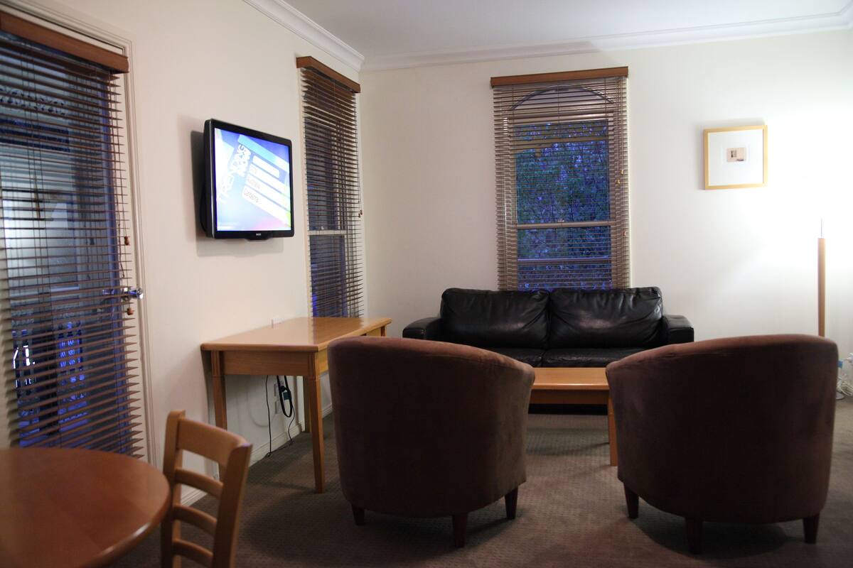 A living room with a black leather couch, two brown armchairs, a wooden coffee table, and a television mounted on the wall.