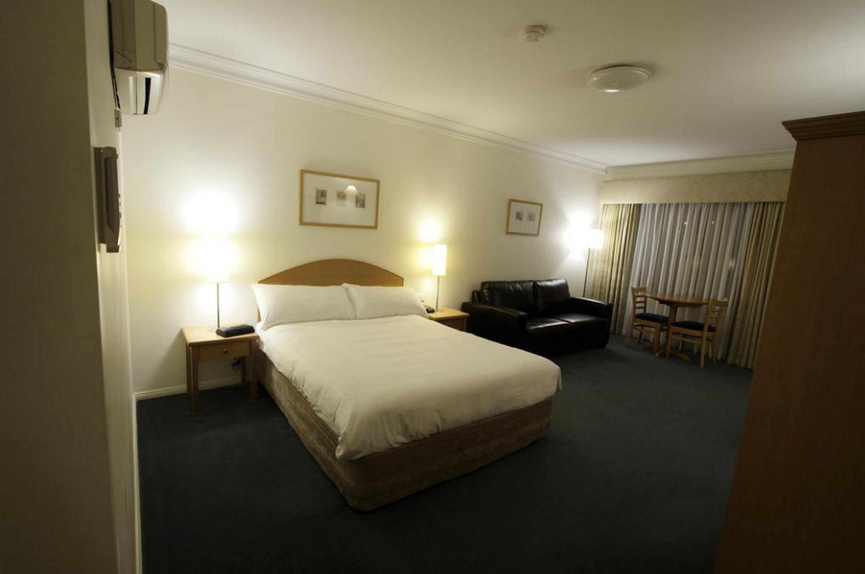 A hotel room with a large bed, two bedside lamps, a leather armchair, a small table, and a view of the outside through curtains.