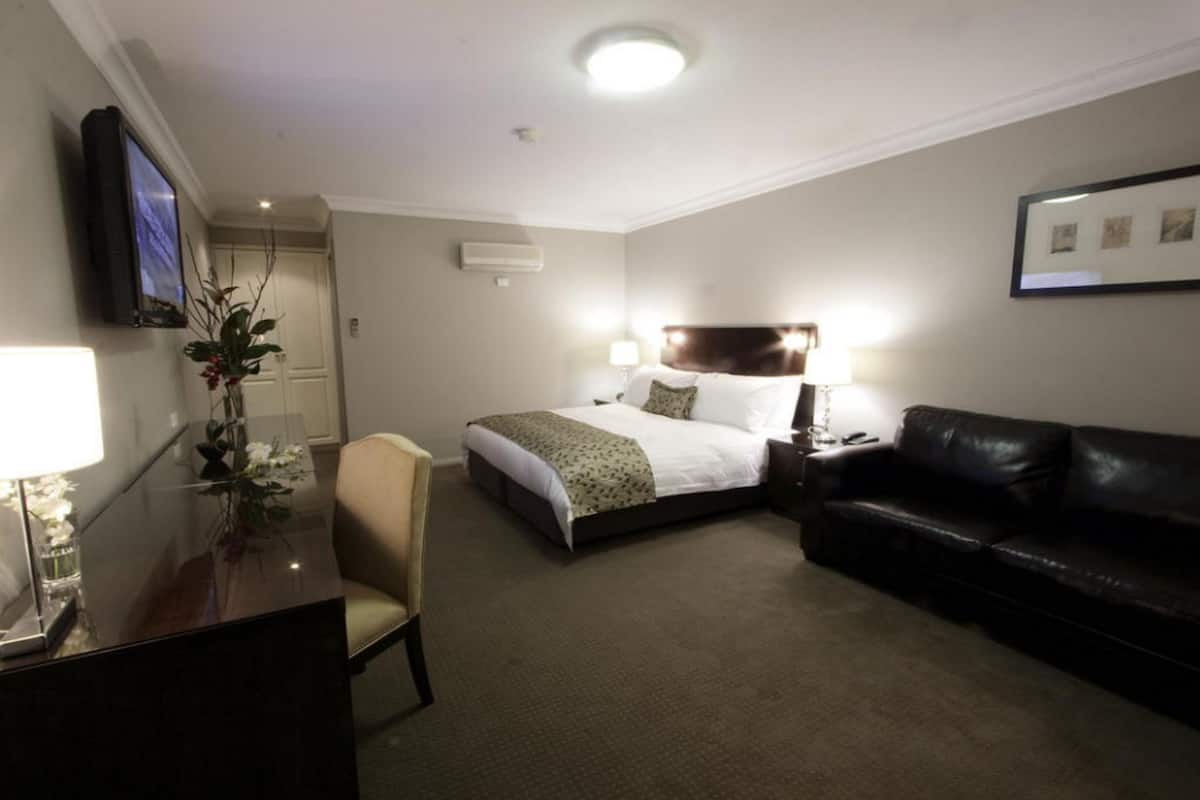 A hotel room with a bed, a desk with a lamp, a chair, a black leather sofa, and a television mounted on the wall.