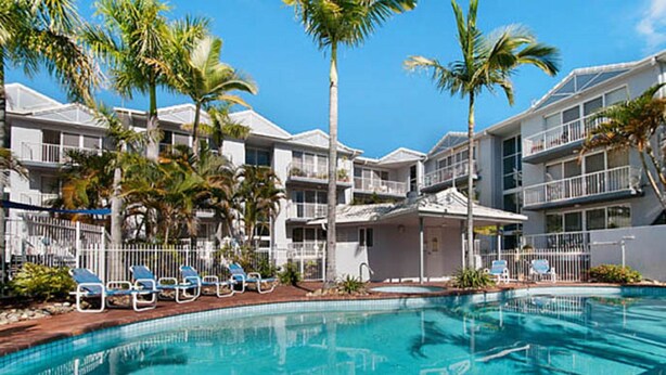 A swimming pool with lounge chairs, palm trees, and a building with balconies. - Champelli Palms (Labrador)