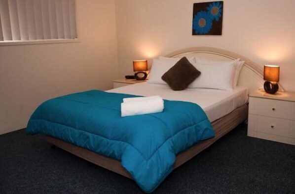 A neatly made bed with a blue comforter and white pillows, two bedside lamps, a small bedside table, and a wall-mounted artwork.