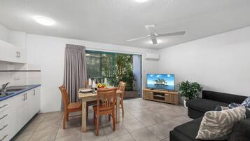 A modern kitchen with white cabinets, a dining table with chairs, a TV, a sofa, and a ceiling fan.