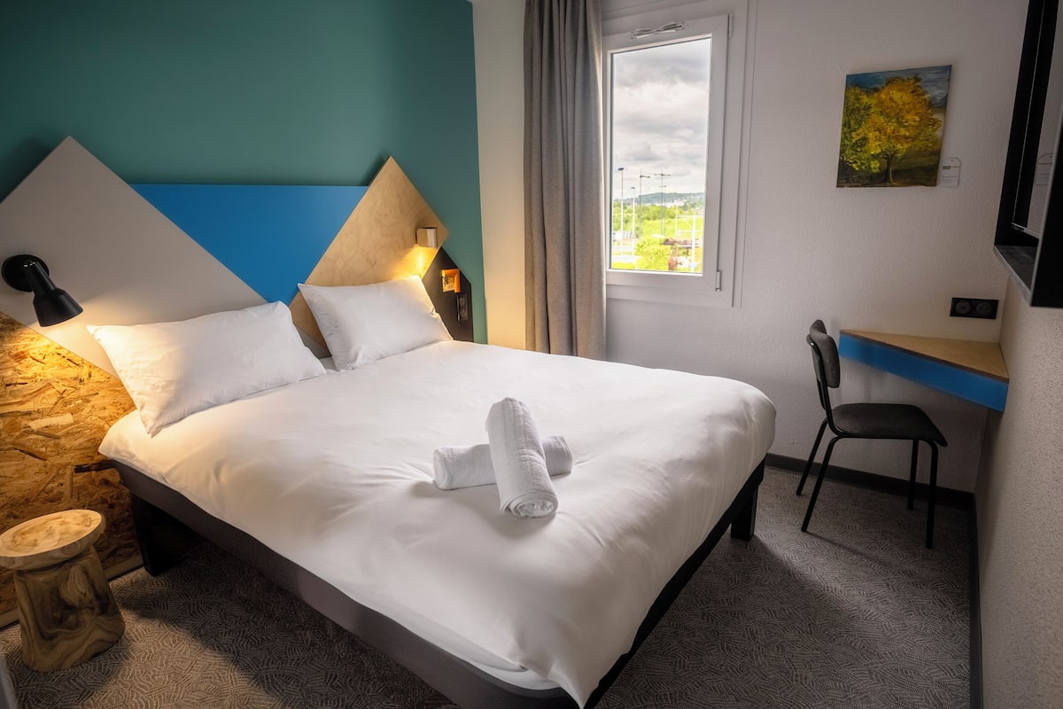 A hotel room with a bed, a desk, a chair, a lamp, and a painting on the wall.