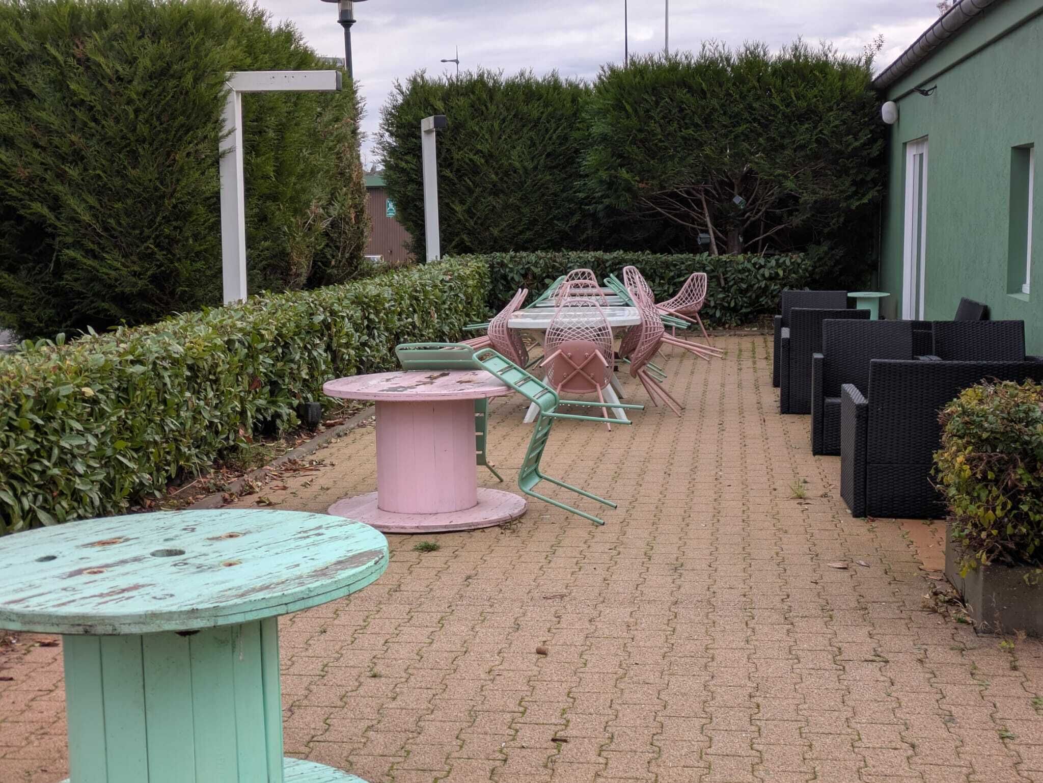 Un patio con mesas y sillas, rodeado de setos y un edificio.
