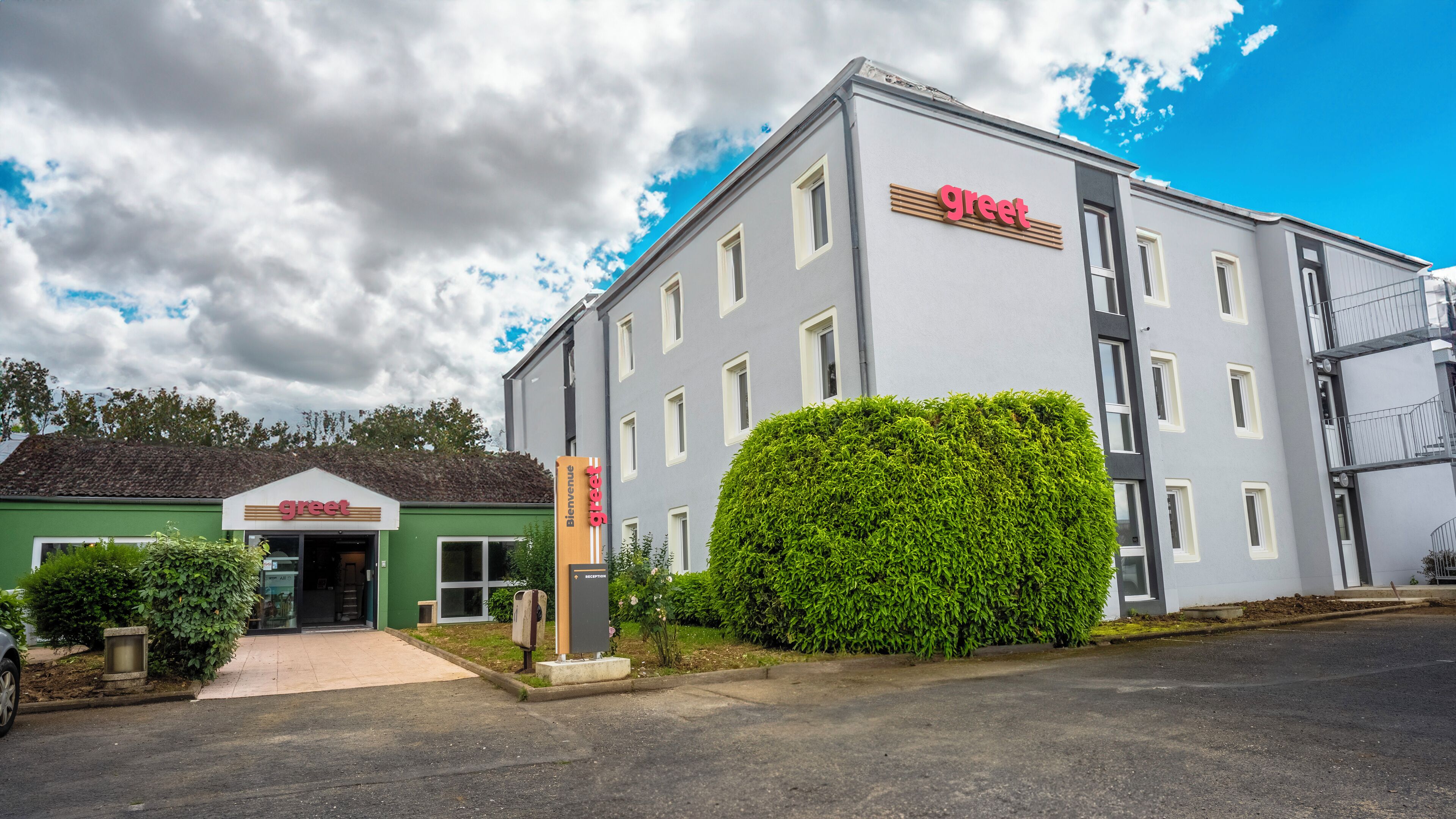 A modern building with a 'greet' sign, a reception desk, and a well-maintained hedge in front.