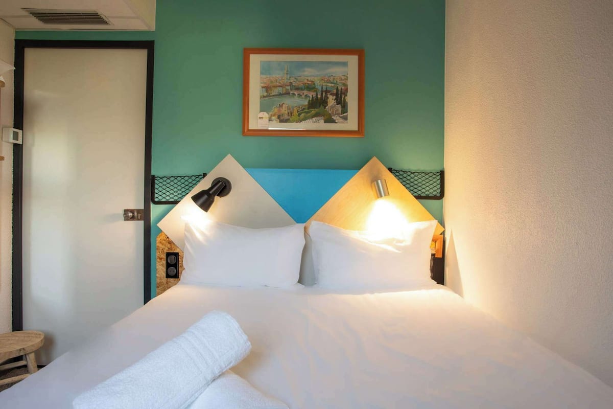 A hotel room with a bed, two pillows, a nightstand, a lamp, a painting on the wall, and a door.