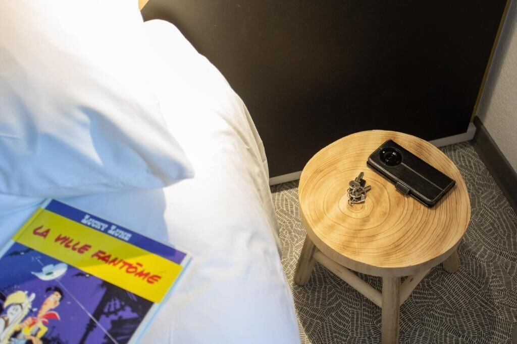 A hotel room with a bed, a small wooden side table with a smartphone and a keychain, and a book titled "LA VILLE FANTOMÉ".
