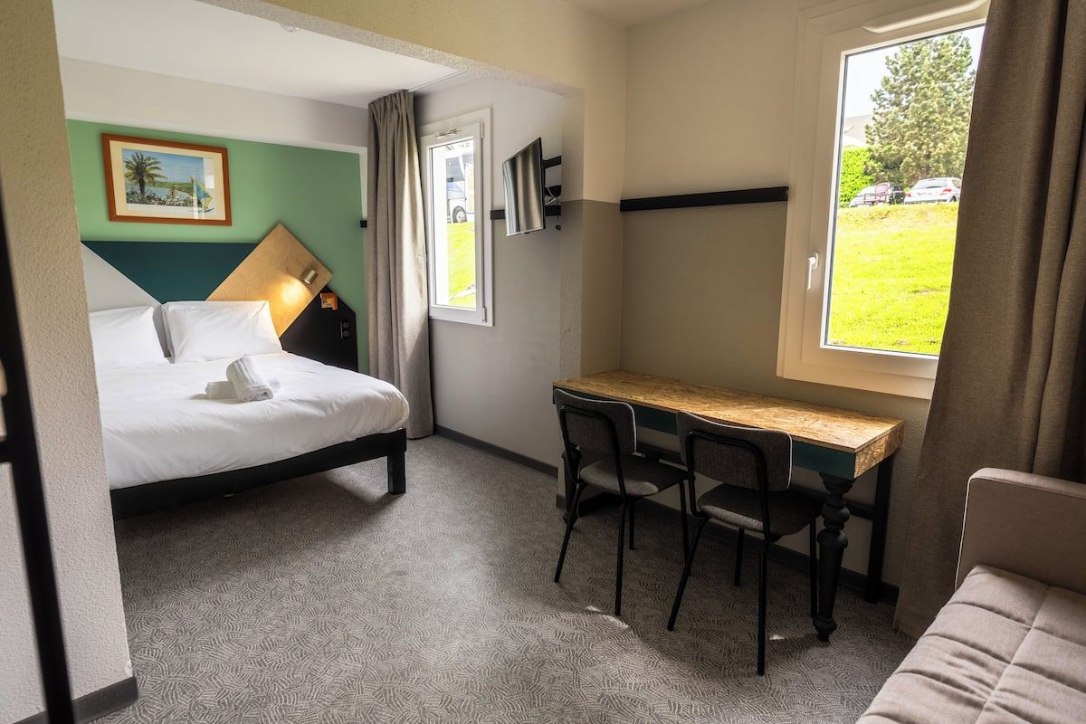 A hotel room with a bed, a desk with two chairs, a window with a view of the outside, and a picture on the wall.