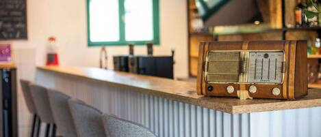 Una zona de bar con una radio antigua, taburetes, un televisor y una ventana con persianas.