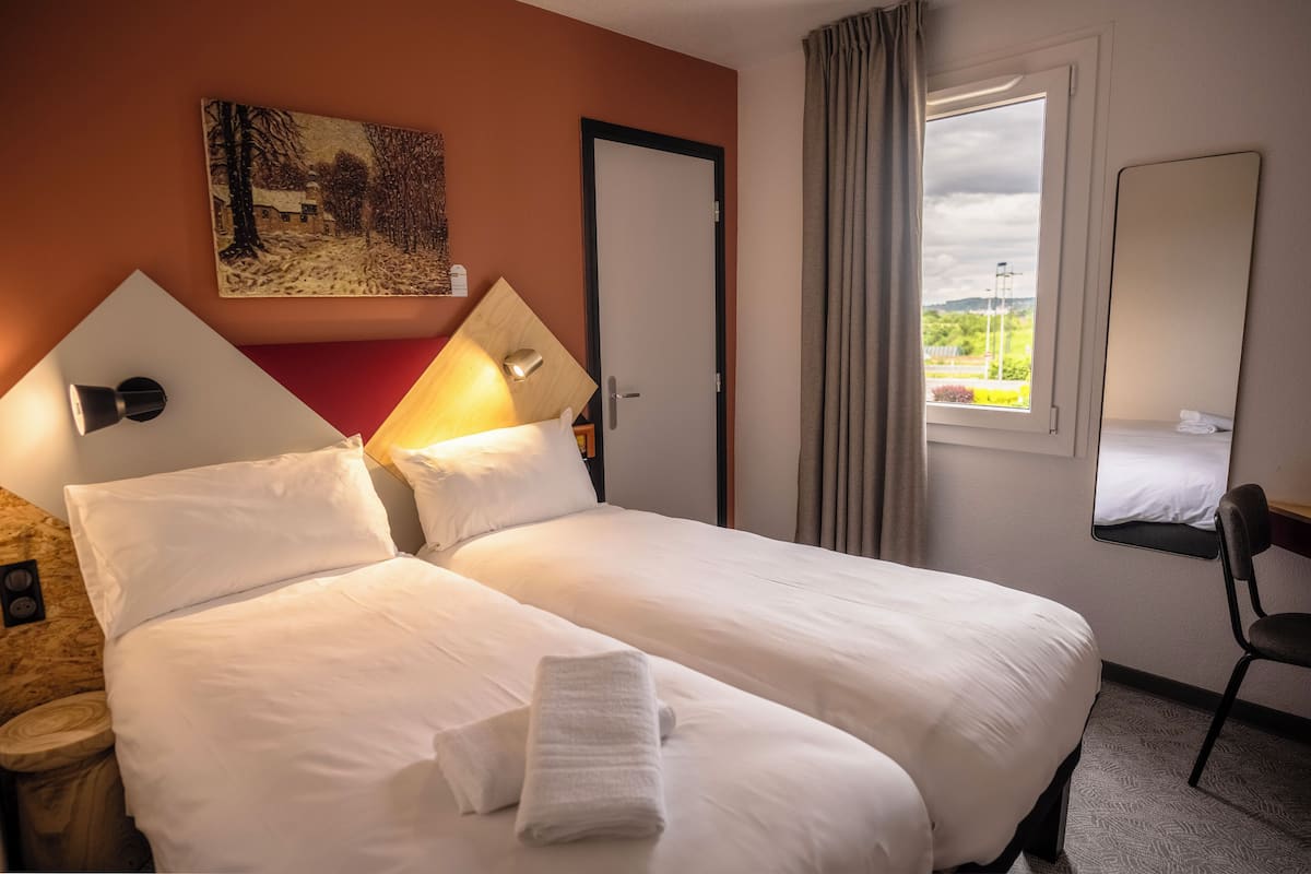 A hotel room with a large bed, a painting on the wall, a window with curtains, and a chair.