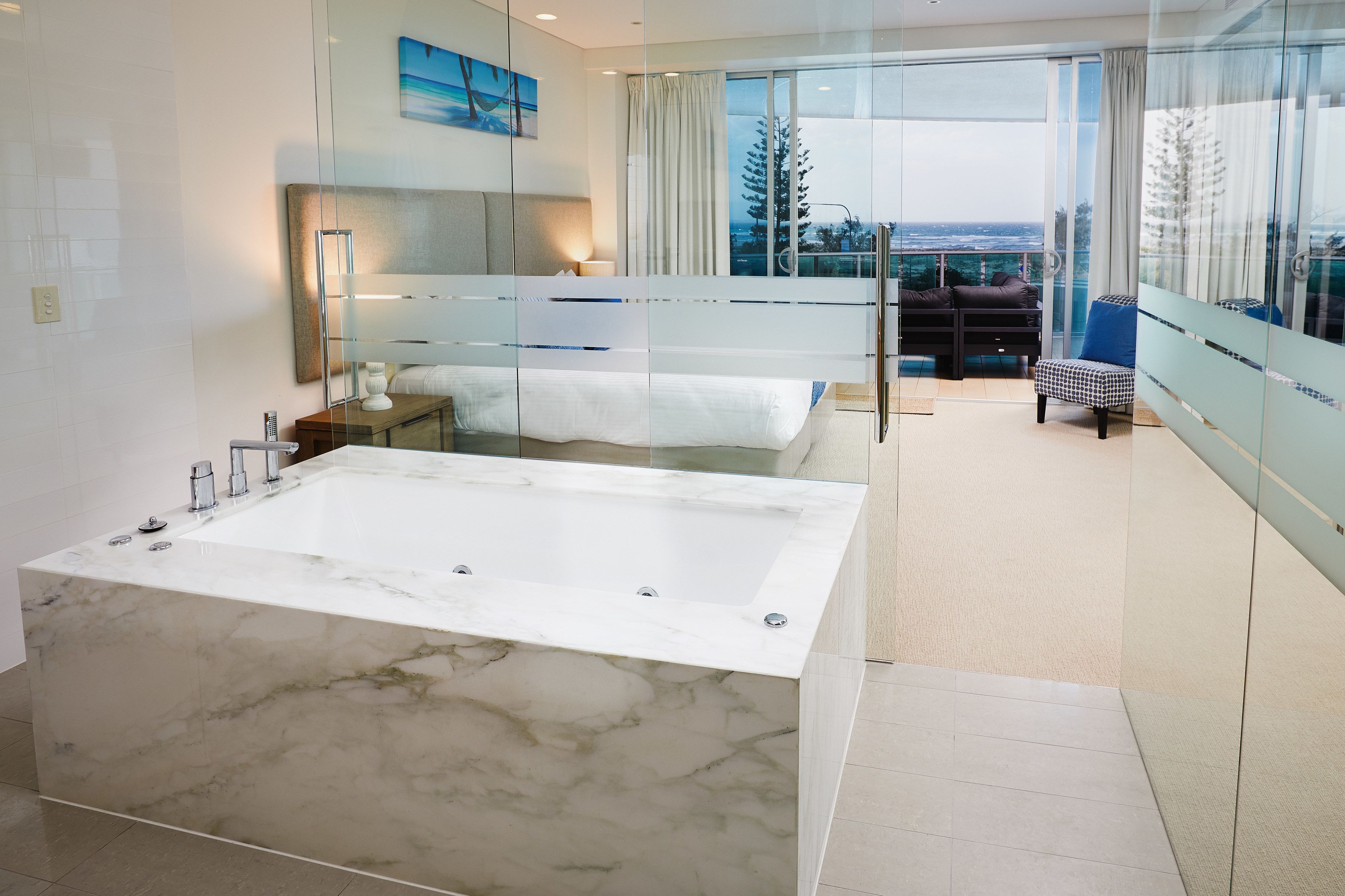 Une salle de bain moderne avec une grande baignoire en marbre, une table de chevet et une commode. Une grande fenêtre offre une vue sur la ville.