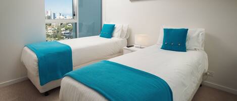 A hotel room with two beds, each with a blue throw blanket, white bedding, and a nightstand with a lamp. There are framed pictures on the wall and a view of the city through a large window.