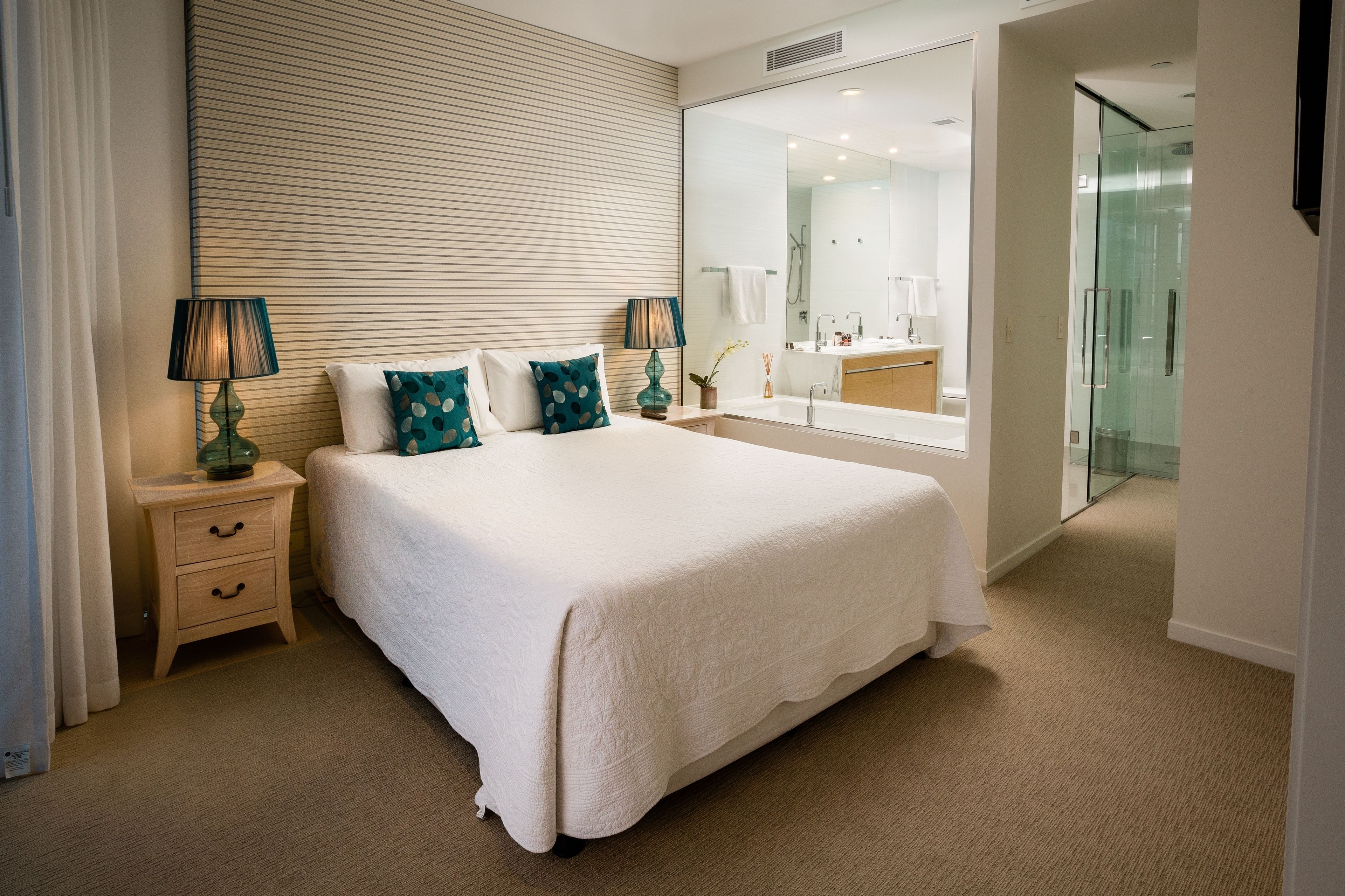 A neatly arranged hotel room with a large bed, two bedside tables with lamps, a mirror, and a glass shower enclosure.