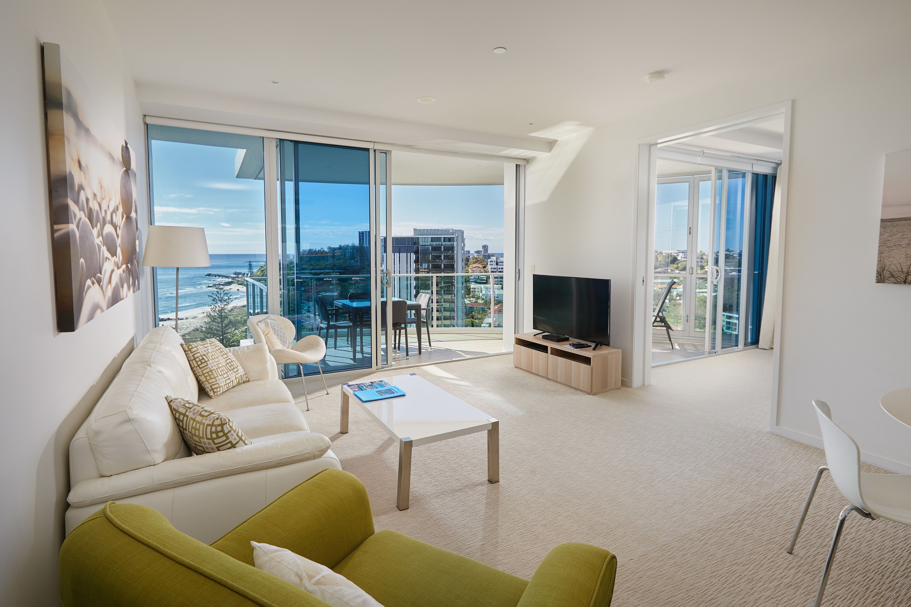 Un salon moderne avec un canapé, un fauteuil vert, une table basse et un téléviseur. De grandes fenêtres offrent une vue sur la ville et donnent accès à un balcon.