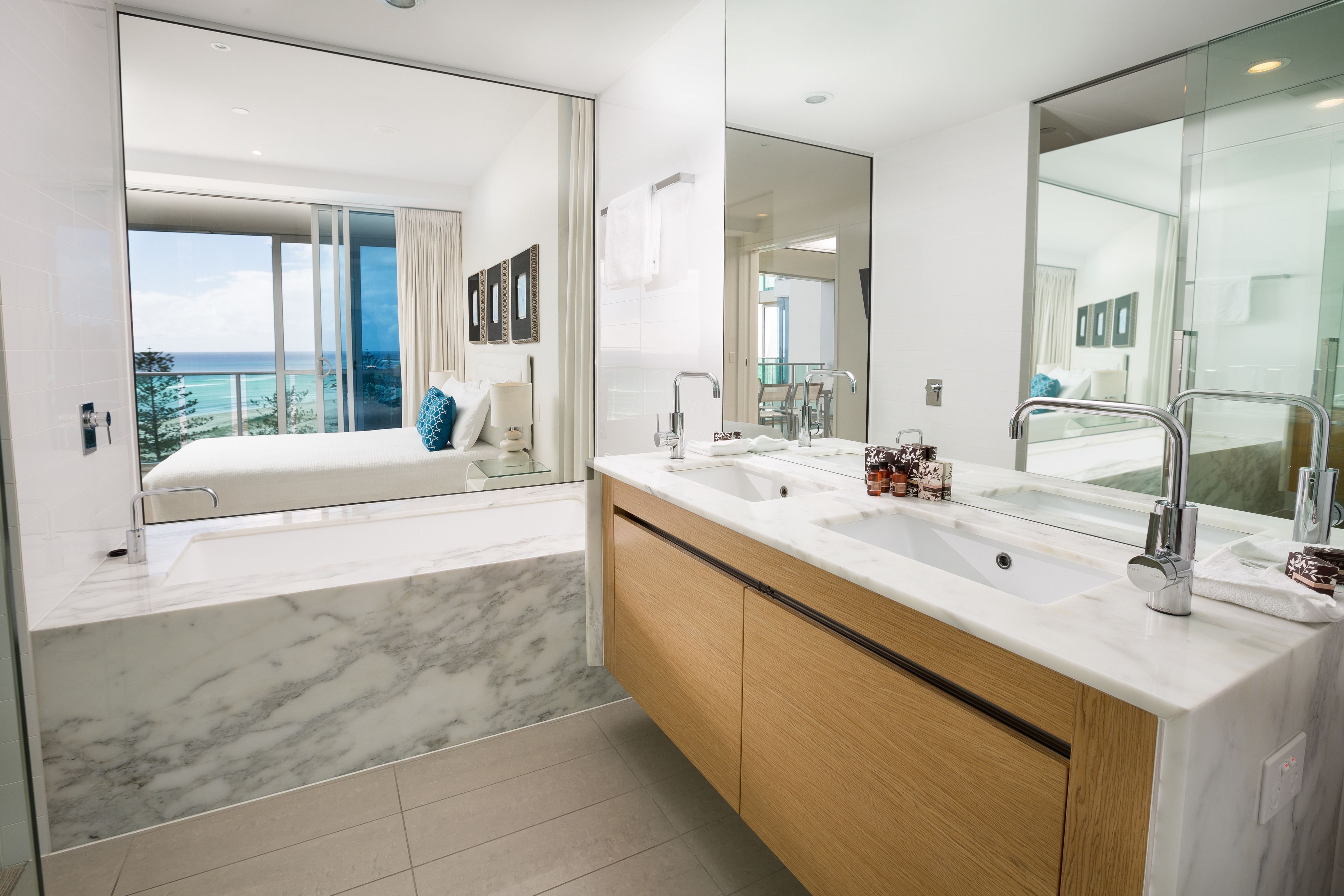 A modern bathroom with a large mirror, a double sink, and a view of a balcony with a bed.