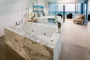 A modern bathroom with a large marble bathtub, a glass-enclosed shower, and a view of the ocean.