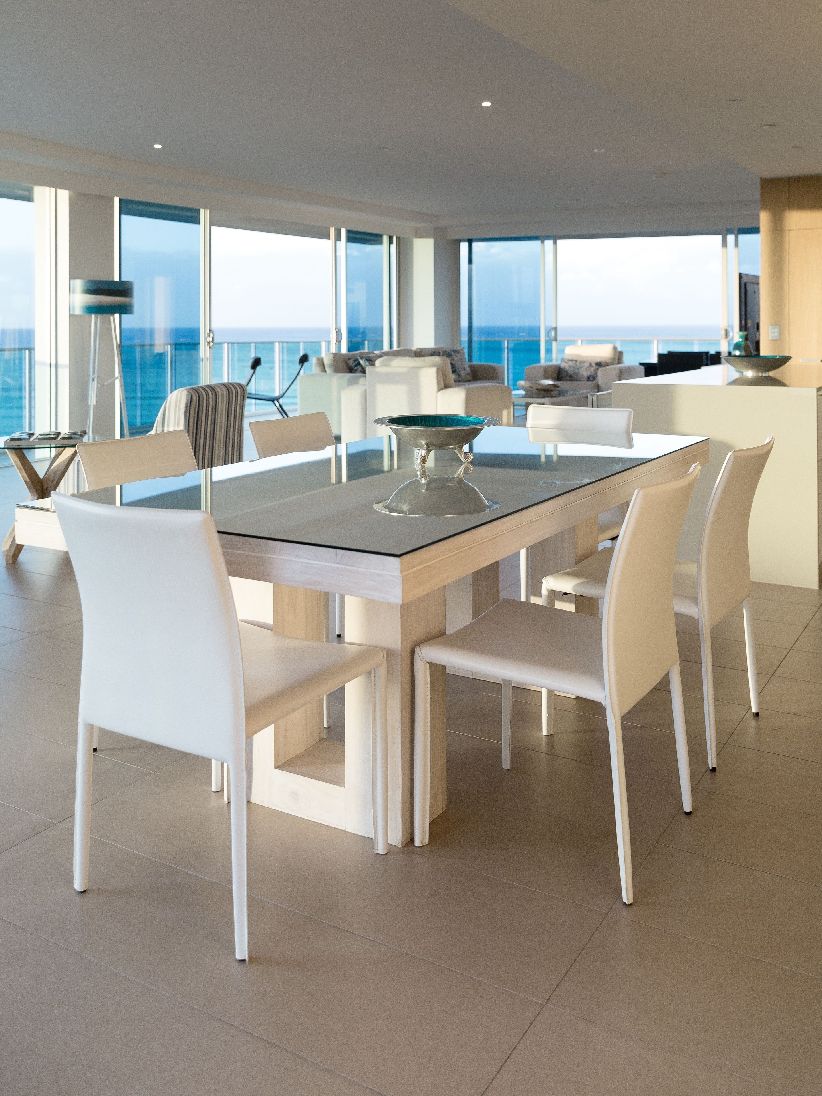 A modern dining area with a glass-top table, white chairs, and a view of the ocean through large windows.