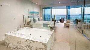 Un baño moderno con una bañera de mármol grande, un balcón con paredes de vidrio y vistas al océano, y un dormitorio con una cama amplia y vista al mar.