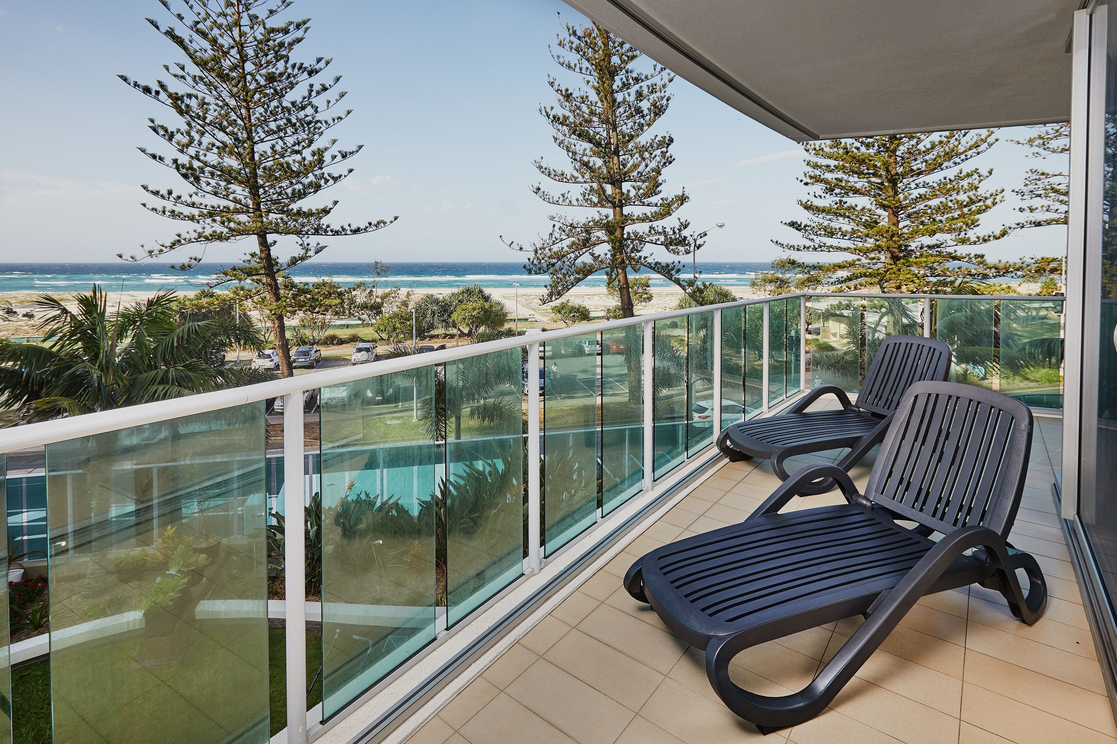 A balcony with two lounge chairs, a glass railing, and a view of the beach and ocean.