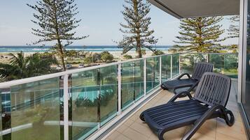 A balcony with two lounge chairs, a glass railing, and a view of the beach and ocean.