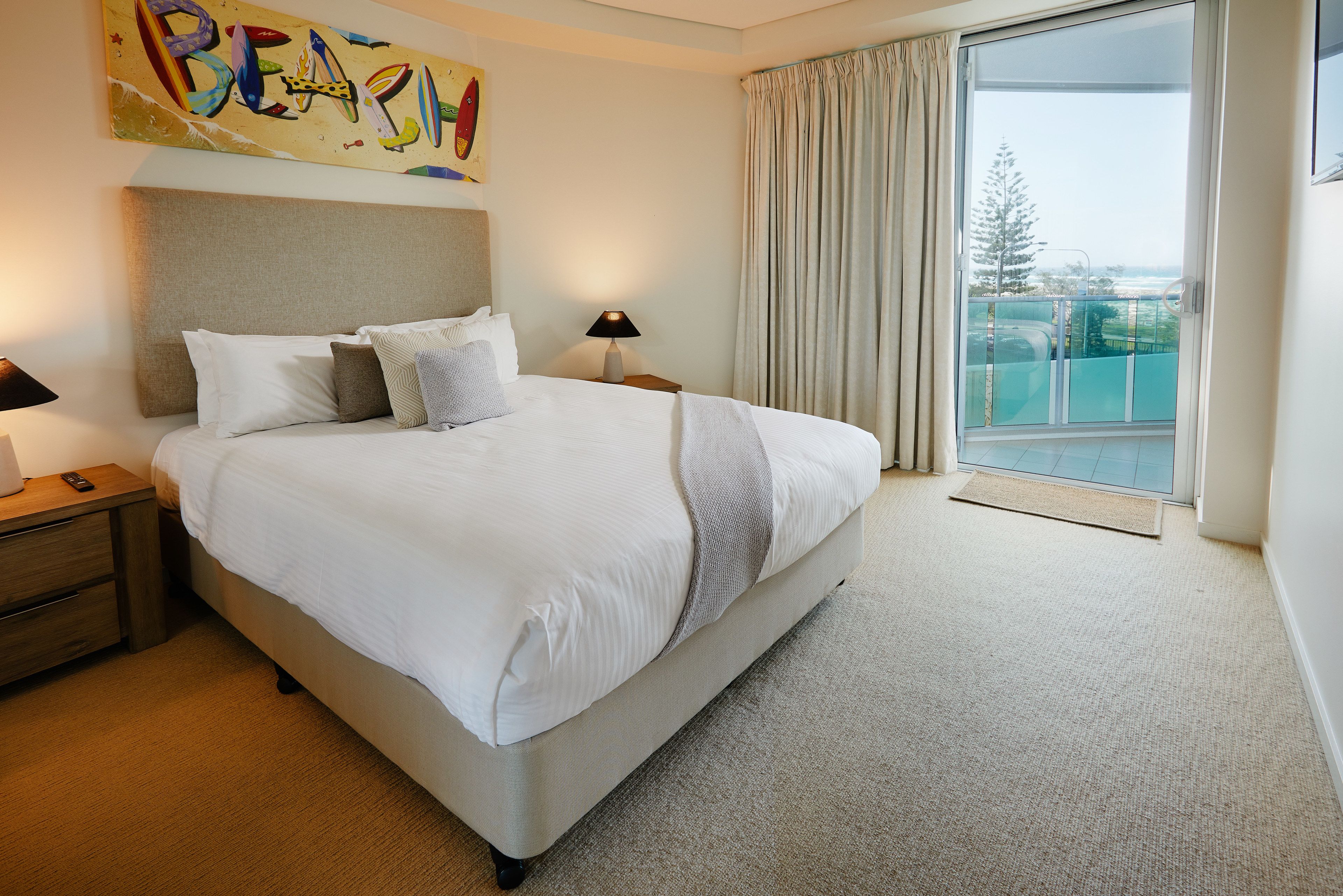 A hotel room with a large bed, a nightstand, a lamp, and a view of the outdoors through a sliding door.