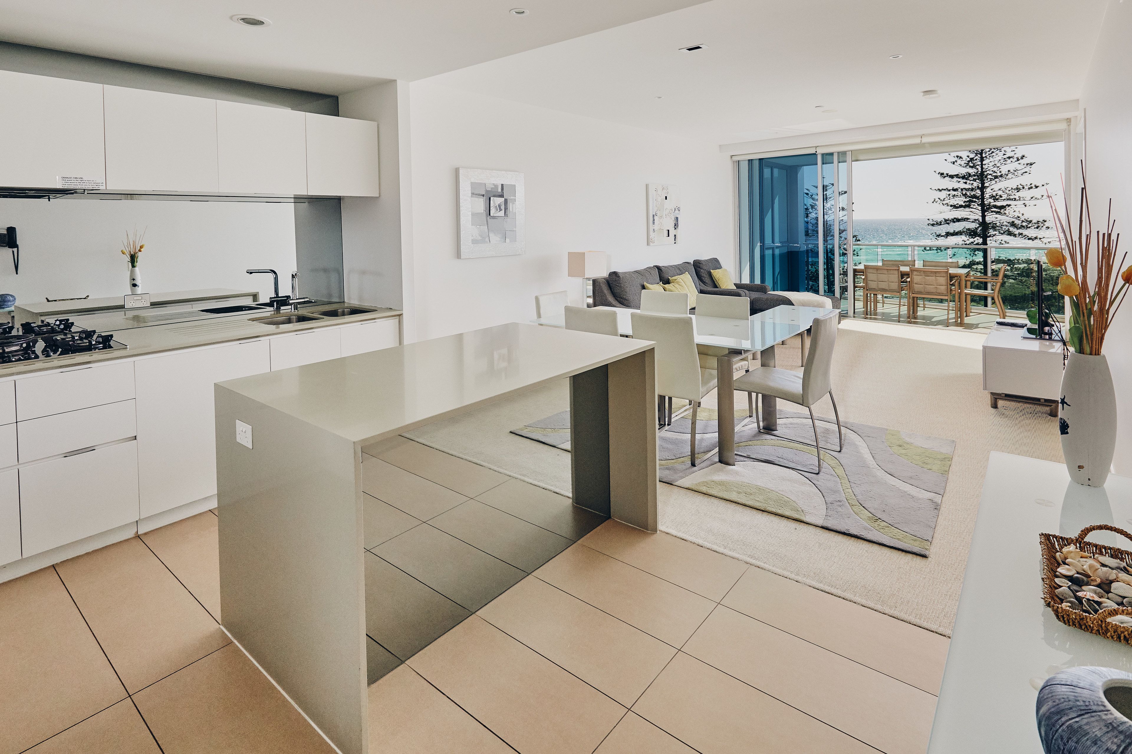A modern kitchen with white cabinets, a dining area with a glass table, and a view of the ocean through large windows.