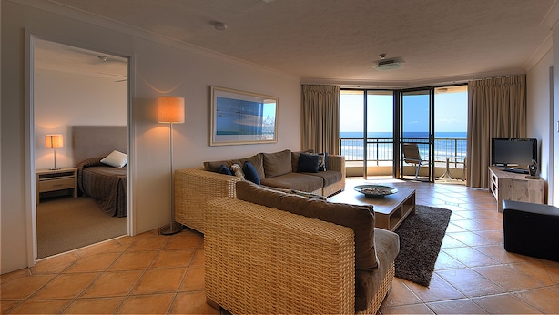 A spacious living room with a large window, a sofa, a coffee table, and a television. - San Simeon Apartments (Tugun)