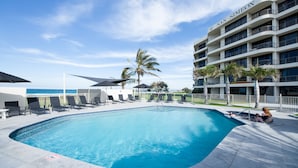 A swimming pool with lounge chairs and umbrellas, a building with balconies, and a palm tree. - San Simeon Apartments (Tugun)