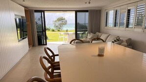 A spacious dining area with a large table and chairs, a television mounted on the wall, and a view of the ocean through large windows. - San Simeon Apartments (Tugun)