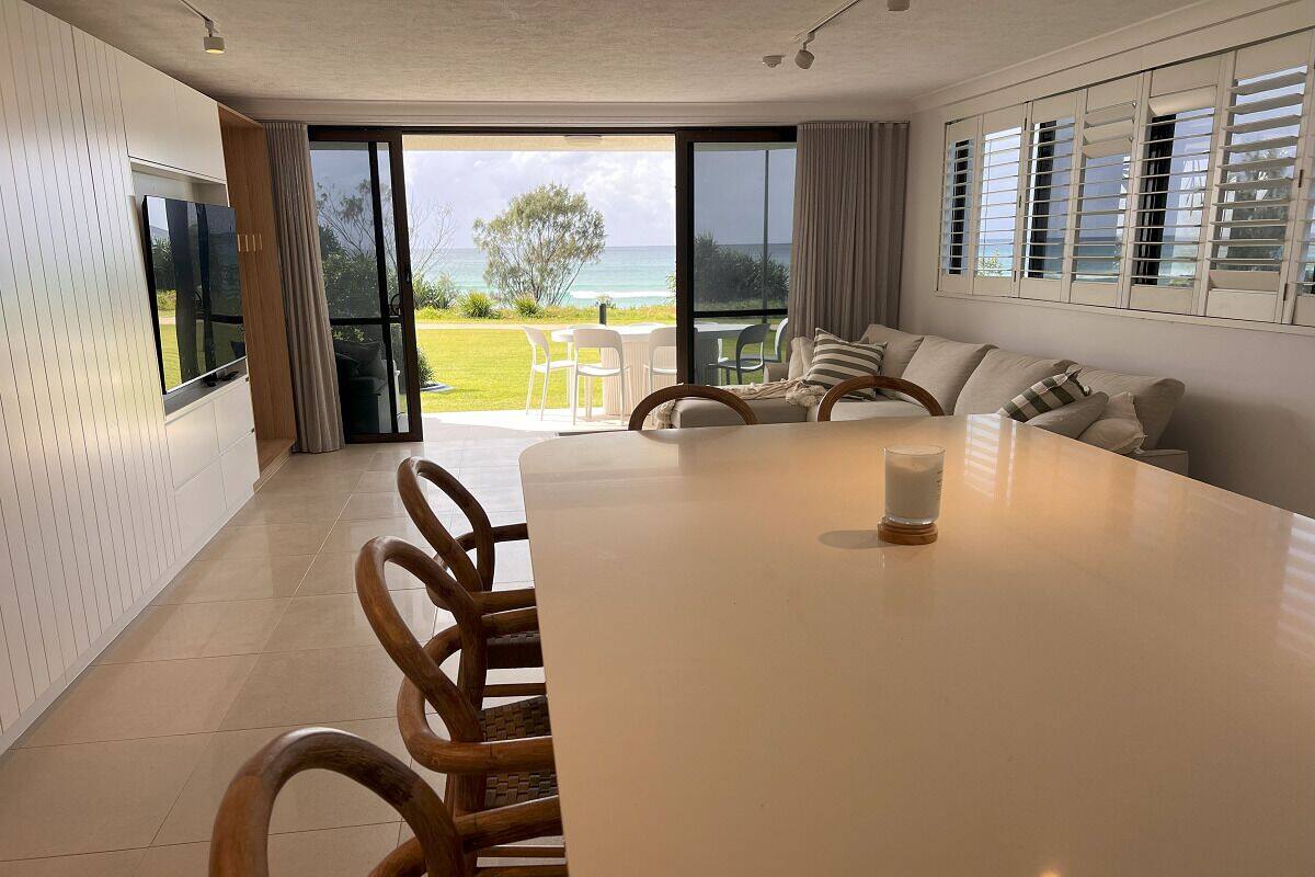 A spacious dining area with a large table and chairs, a television mounted on the wall, and a view of the ocean through large windows.