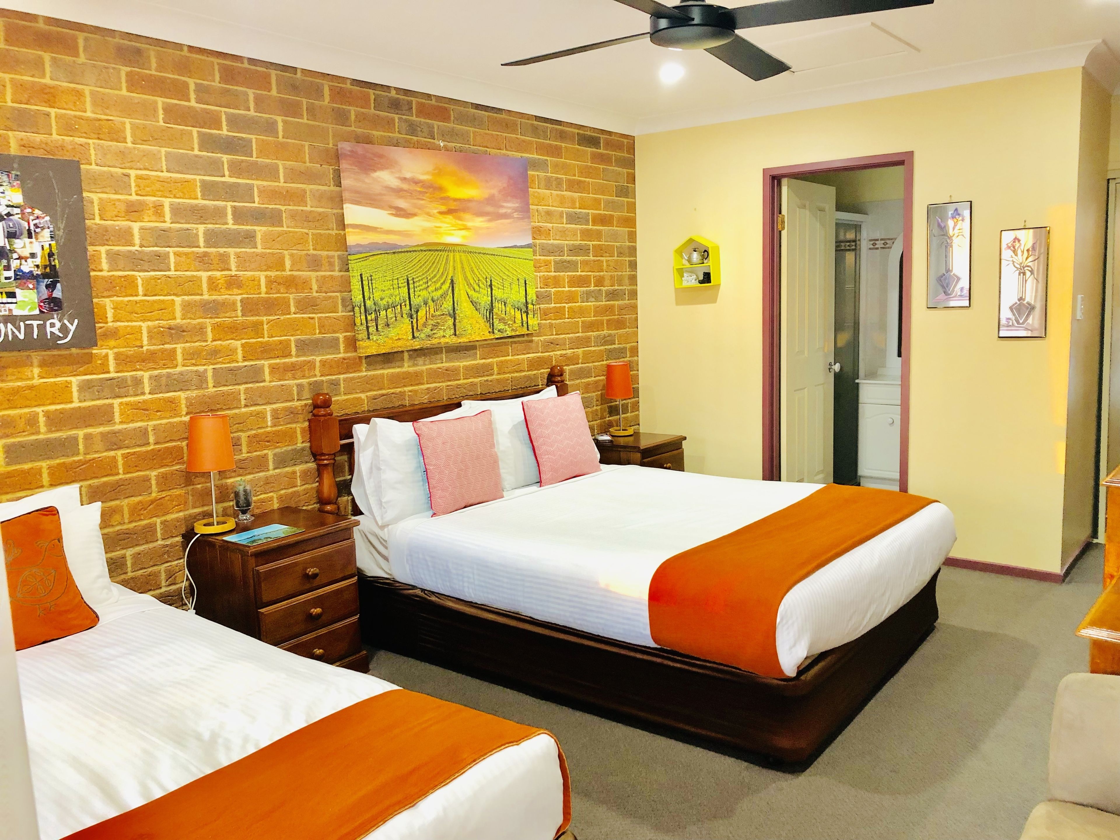 A hotel room with two beds, a brick wall, a ceiling fan, and a painting of a vineyard.