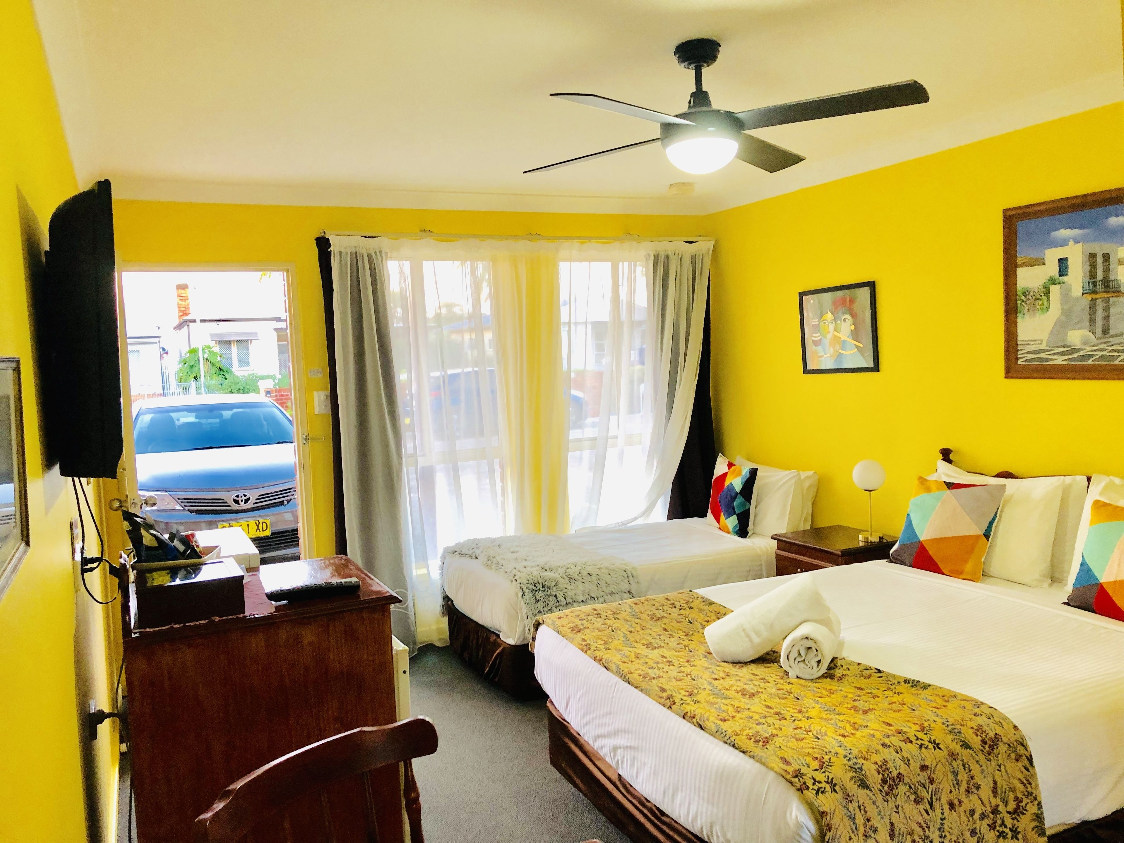 A hotel room with two beds, a desk, a ceiling fan, and a view of the street outside.
